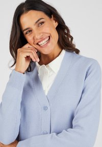 Cardigan a maglia azzurro chiaro con scollo a V, tre bottoni e polsini a costine. Indossato sopra una camicia bianca con colletto, caratterizzata da una texture liscia.