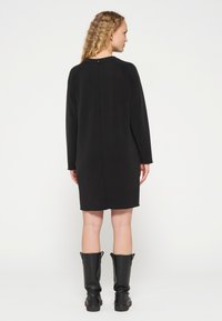 Vestido negro de manga larga con un corte relajado, que presenta un escote redondeado y un detalle de costura en la parte posterior. Combinado con botas de cuero negras hasta la rodilla.