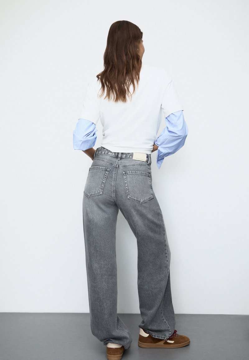 Jean gris à jambes larges avec une taille haute, doté de poches arrière et d'une étiquette. Associé à une chemise blanche à rayures bleues, style décontracté.