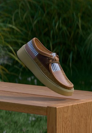 Casual bootschoen met bruin leer, blauw-wit gestreepte stof en een beige zool, zwevend boven een houten bankje buiten.