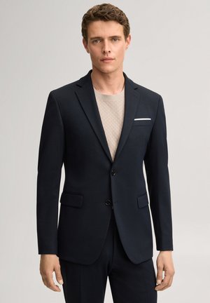 Man draagt een aansluitend zwart colbert over een beige textuurachtig overhemd, staand tegen een effen lichtgrijze achtergrond.