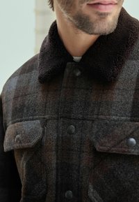 Close-up di un uomo che indossa una giacca di lana a quadri scuri con tasche a patta e un colletto testurizzato in stile shearling.