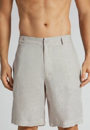 Lyse beige knælange hørshorts båret af en person uden skjorte med arme afslappet langs siden mod en ensfarvet baggrund.