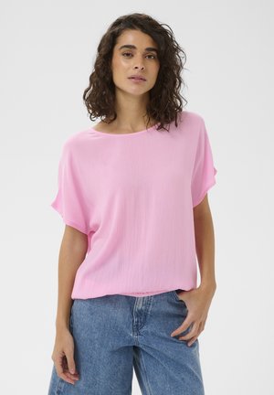 Woman with curly brown hair wearing a loose pink short-sleeve top and blue denim jeans, standing with one hand in pocket.