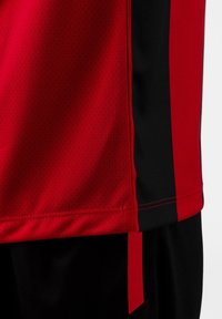 Maglietta sportiva rossa e nera realizzata in tessuto traspirante, con texture perforata e pannelli di colore contrastante lungo i lati.