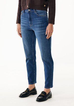 Frau in mittelhohen, enganliegenden blauen Jeans, schwarzen Leder-Loafers und einer dunkelbraunen, cropped Strickjacke vor weißem Hintergrund.