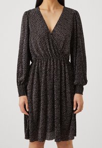 Kvinna iklädd en svart klänning med beige prickmönster, långa puffärmar, v-ringad halsringning, elastisk midja och volangkjol.