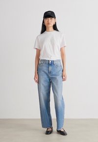 Bijela pamučna majica s grafičkim detaljima, svijetloplave traperice visokog struka, crna kapa i crne ravne cipele. Jednostavan, le�žeran outfit.