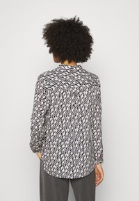 Camicia a maniche lunghe in tessuto stampato con uno sfondo bianco e forme a diamante blu e nere, colletto e orlo arrotondato.