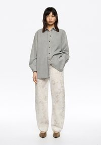 Chemise boutonnée surdimensionnée gris clair avec une texture douce, assortie à un pantalon fluide à motifs blanc cassé. Des chaussures marron complètent la tenue.