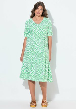 Grünes, gemustertes Kleid mit V-Ausschnitt und kurzen Ärmeln. Verfügt über ein fließendes, gestuftes Design. Getragen mit braunen Sandalen.