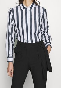 Knapp-up skjorta med blå och vita vertikala ränder, långa ärmar, krage och en bröstficka, ihop med högmidjade svarta byxor.