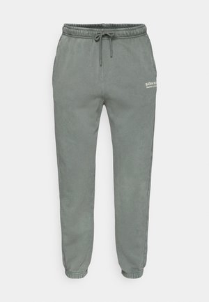 Grå joggebukser laget av mykt stoff, med elastisk liv med snøring, avsmalnede ankler og en logo trykt på venstre lår.