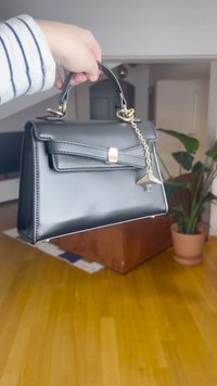 Fekete műbőr kézitáska strukturált kialakítással, felső fogantyúval és arany színű láncos díszítéssel. Masnis formájú fedél és kapcsos zár jellemzi.