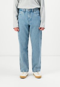 Cilvēks ar gaiši ziliem taisnām biksēm, pelēku garām piedurknēm kreklu un baltām sporta apavām ar gumijas zoli stāv pret baltu fonu.