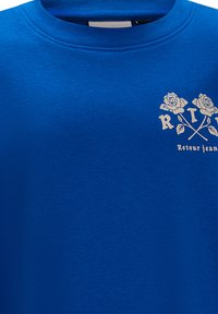 Felpa in cotone blu con grafica di rose e frecce incrociate, accompagnata dal testo "R I Retour jeans" in bianco. Texture liscia e scollatura rotonda.