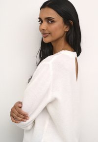 Hvid, blød striktrøje med rund halsudskæring, der har et åbent rygdesign. Stoffet virker fluffy med en afslappet pasform.