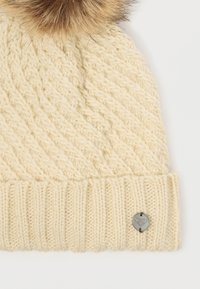 Knit cream beanie with a textured pattern, featuring a large faux-fur pom-pom on top and a round metal accent on the cuff.