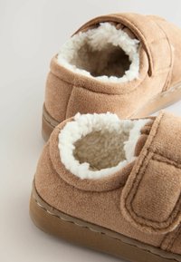 Barna suede papucsok puha fehér gyapjú bélelésse, hátsó rész nélküli kialakítás és tépőzáras pánt a könnyű felvétel érdekében. Texturált talp a tapadásért.