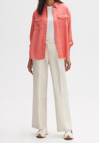 Korallenfarbenes Hemd mit Knopfleiste und zwei Brusttaschen, kombiniert mit cremefarbenen Hosen mit weitem Bein. Die Sneakers zeigen eine Mischung aus Beige-, Creme- und Blautönen.