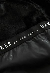 Tissu en fausse fourrure noire avec une texture lisse, doté d'une étiquette noire portant l'inscription "by Ted Baker" en lettres blanches.