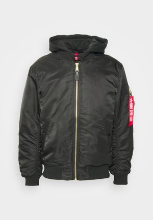 Blouson noir avec capuche, fermeture éclair, poignets côtelés, poches latérales et un détail en étiquette rouge sur la manche gauche. Tissu lisse et texturé.