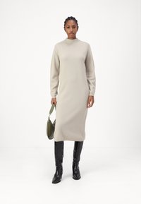 Robe tricotée beige clair avec un col montant et des manches longues, présentant une silhouette ajustée. Le mannequin tient un sac à main vert foncé, portant des bottes noires jusqu'aux genoux.
