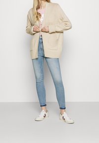 Cárdigan de punto beige con un ajuste relajado, grandes bolsillos y puños acanalados, combinado con jeans ajustados de color azul claro y zapatillas blancas.