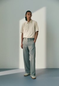 Lichtbeige korte mouwen knoopshirt gecombineerd met lichtgrijze wijde broek en witte sneakers, geplaatst tegen een zachte, minimalistische achtergrond.