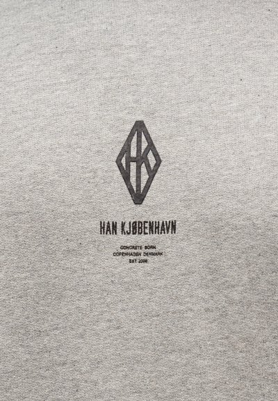 Γκρι ύφασμα με μαύρο γεωμετρικό λογότυπο και κείμενο "HAN KJØBENHAVN" και μικρότερο κείμενο που αναφέρεται στην Κοπεγχάγη της Δανίας και την ίδρυση το 2008.