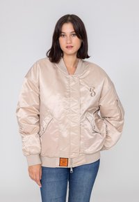 Jasnobeżowa satynowa kurtka bomberka o luźnym kroju, z prążkowanymi mankietami i pasem, kieszeniami na zamek oraz pomarańczową metką u dołu.