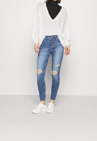 Vit överdimensionerad tröja med V-ringning, ihopparad med blå rivna skinny jeans med knappgylf, och vita sneakers.
