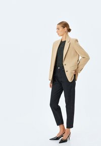 Beige skräddarsydd kavaj, svart texturerad topp, mörka jeansbyxor, svarta lackskor. Enkel design, strukturerad form och minimalt med detaljer.