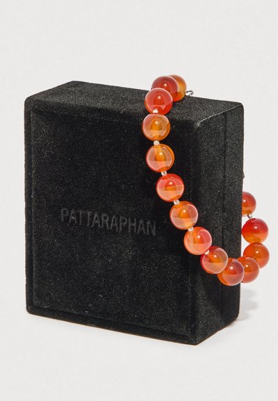 Βραχιόλι με κόκκινες-πορτοκαλί χάντρες τοποθετημένο πάνω σε μια μαύρη βελούδινη θήκη κοσμημάτων με ανάγλυφη την επωνυμία Pattaraphan.