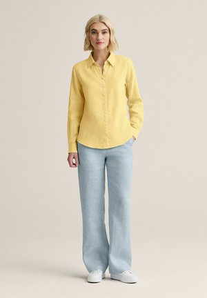 Kvinde står iført en langærmet gul skjorte med knapper, lyseblå bukser med vide ben og hvide sneakers mod en ensfarvet baggrund.