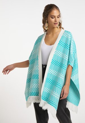 Donna che indossa uno scialle turchese e bianco con motivo a spina di pesce e frange, top bianco, pantaloni neri e orecchini colorati.