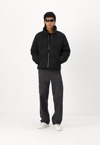 Mustric puffer jakk, millel on lukk, õmblustega õlavarreosad ja soonikkoes mansetid; kombineeritud tumehallide cargo-pükstega ja valgete tossudega. Mustad päikeseprillid on peas.