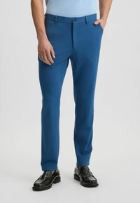 Blauwe op maat gemaakte broek van een gladde stof met een slanke pasvorm. Voorzien van een rits en knoopsluiting aan de voorkant, met zijzakken. Draagt met zwarte schoenen.