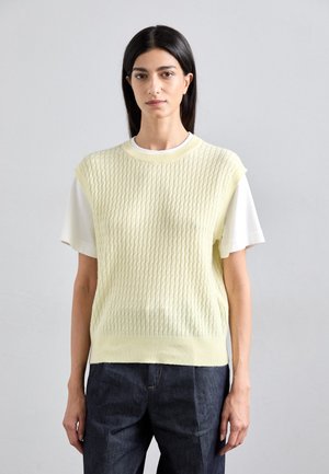 Lys gul strikket genser med teksturert ribbing, kortermet over en hvit skjorte. Kombinert med mørk denim jeans. Enkel og moderne design.