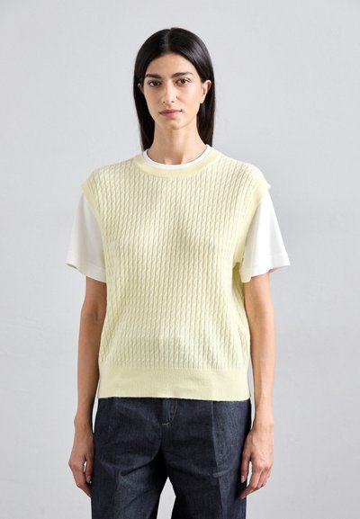 Un pull en tricot jaune clair avec un relief texturé, à manches courtes, sur une chemise blanche. Assorti à un jean en denim foncé. Design simple et moderne.