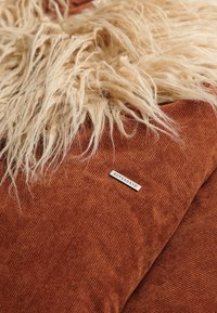 Roodbruine pluche deken met lange, zachte, crèmekleurige nepbont rand en subtiele ribbelstructuur. Bevat een klein merklabel.