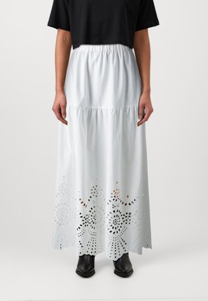Kvinde iført en hvid maxinederdel med hulmønstre ved kanten, kombineret med en sort cropped top og sorte ankelstøvler.