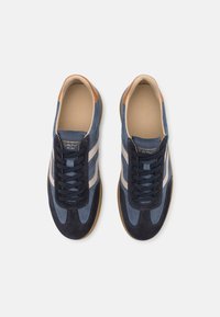 Zapatillas de gamuza azul con rayas blancas, acentos de cuero marrón y cordones oscuros. Punta redondeada y suela de goma texturada. Diseño atlético clásico.