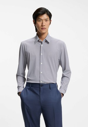 Man wearing a patterned long-sleeve button-up shirt tucked into navy blue dress pants, standing with hands in pockets.