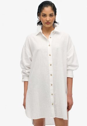 Abito camicia bianco in lino con chiusura a bottoni, maniche lunghe e vestibilità ampia. Presenta un colletto e una texture sottile.