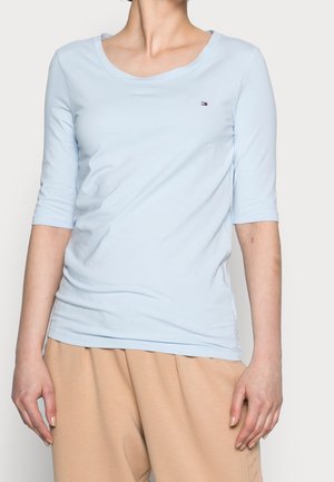 Femme portant une chemise bleu clair à manches trois-quarts avec un petit logo sur la poitrine et un pantalon ample beige, debout devant un fond blanc.