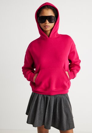 Person iført en lys rosa hettegenser med hendene i lommene, store svarte solbriller, og et grått plissert skjørt med nålestriper.