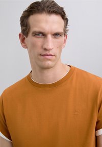 Bărbat cu păr scurt, creț, de culoare brunetă și ochi albaștri, purtând un tricou portocaliu cu manșete albe răsucite, îndreptându-se spre camera foto pe un fond gri simplu.