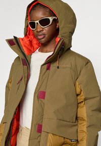 Oliwkowo-zielona ocieplana kurtka z pomarańczową podszewką, kontrastującymi bordowymi wstawkami, kapturem oraz wieloma detalami zapinanymi.