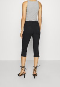 Mujer vestida con pantalones cortos negros, top sin mangas gris y sandalias de tacón alto negras con tiras, de pie contra un fondo liso.